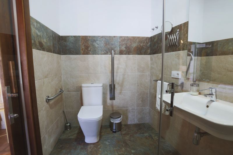Baño adaptado con ducha a ras de suelo, barras de apoyo junto al inodoro y lavabo sin mueble