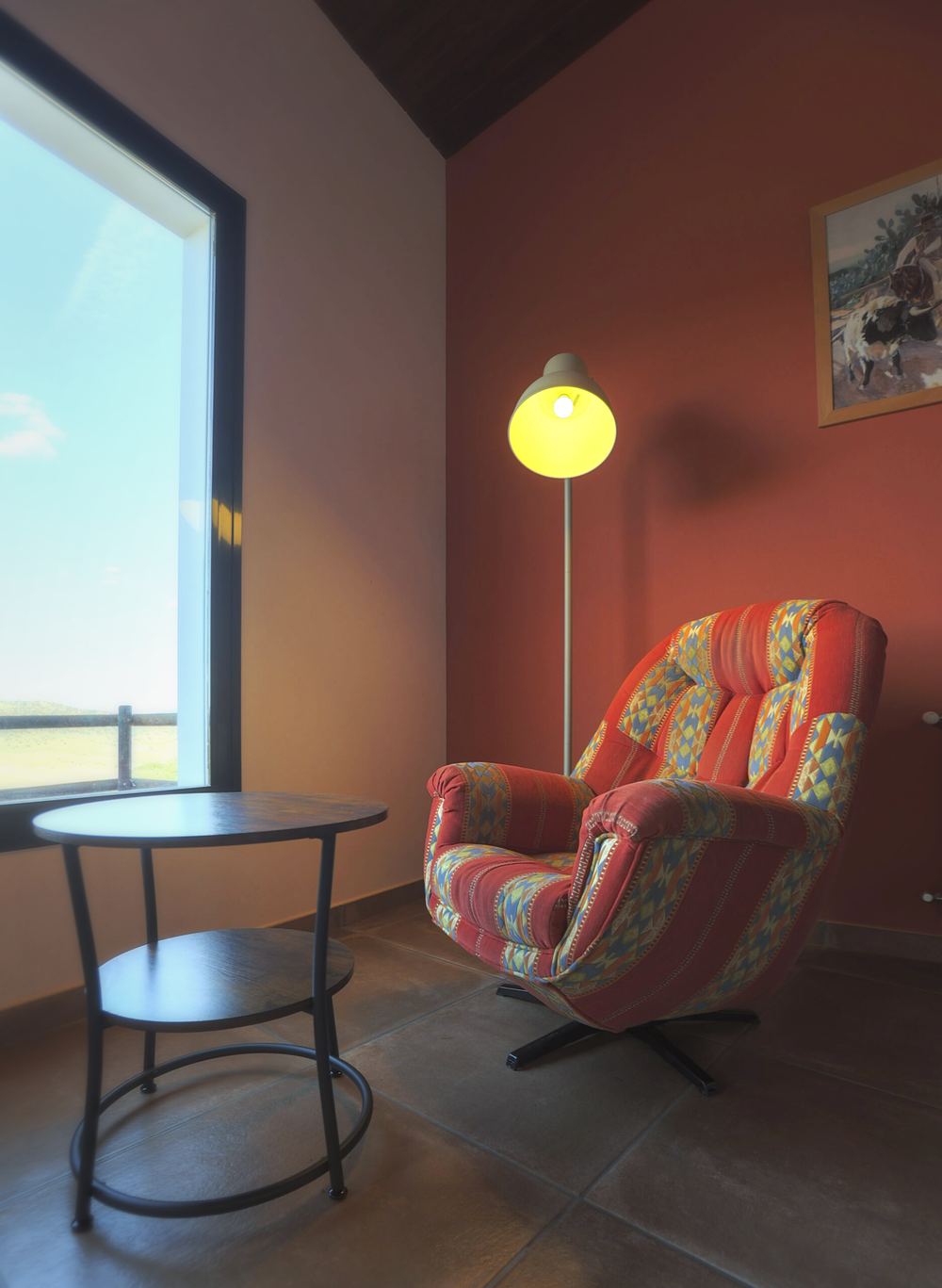 Rincón de una habitación con pared de almagre, una butaca de tela de colores, una lámpara de pie y una ventana grande al campo
