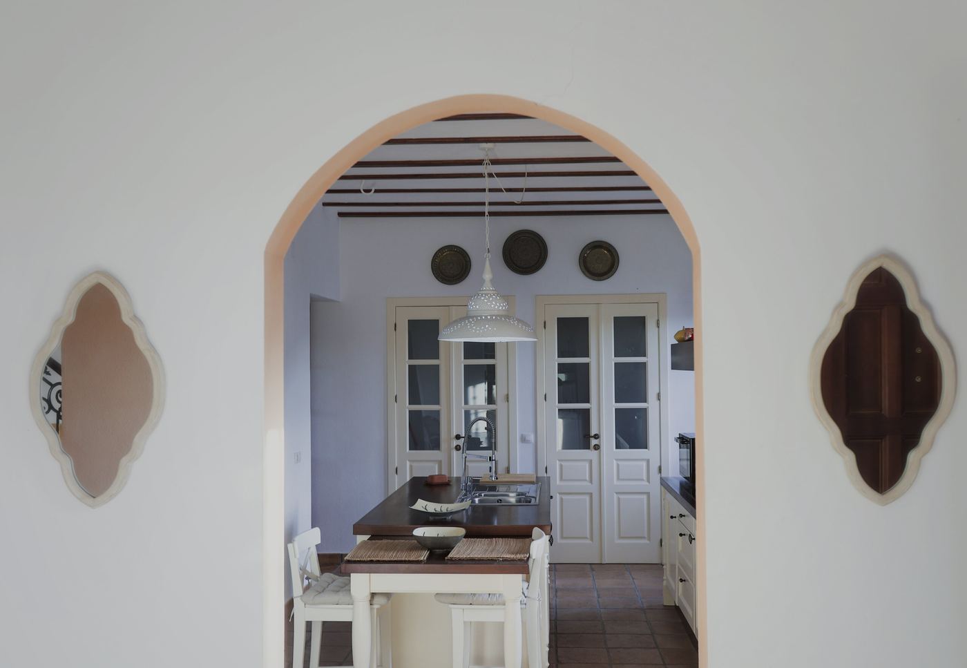 Cocina vista a través de un vano encalado, con isla de madera, taburetes blancos y vigas en el techo