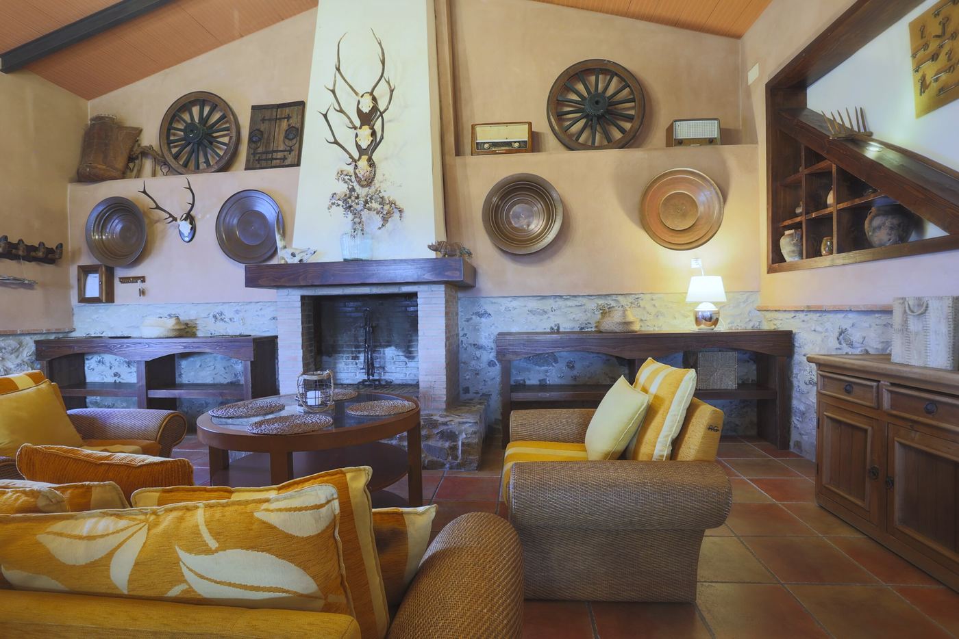 Salón con chimenea, sofás de mimbre con cojines amarillos, ruedas de carro y bandejas de cobre en la pared