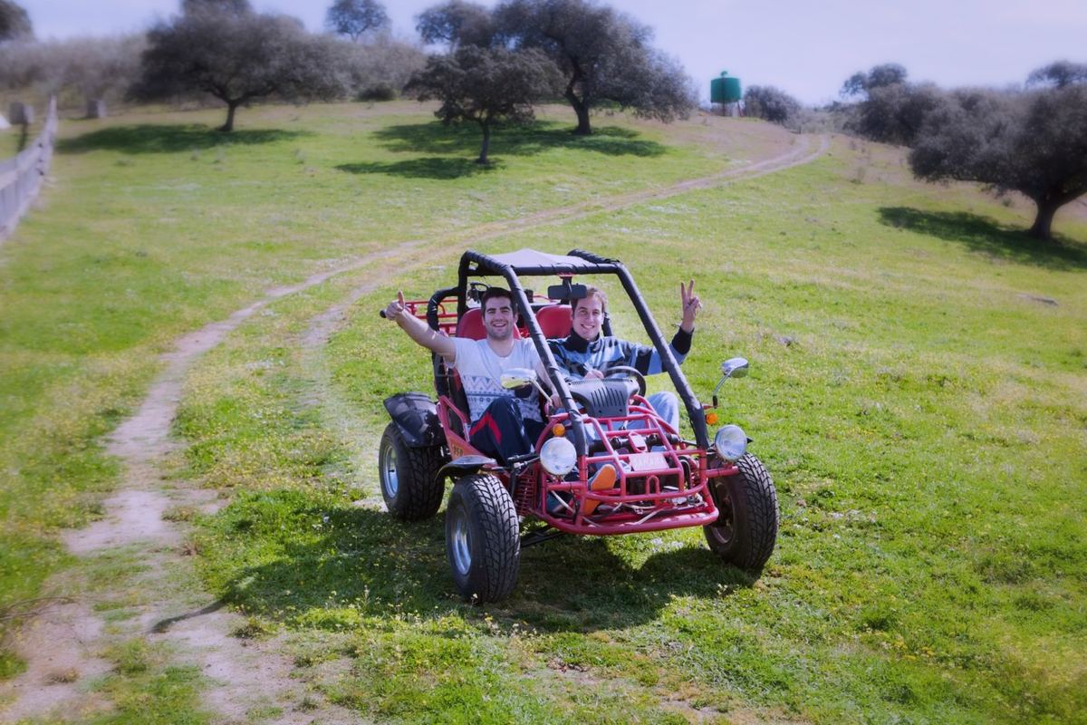 Dos chicos saludan desde un buggy rojo en un camino de tierra entre la hierba y las encinas