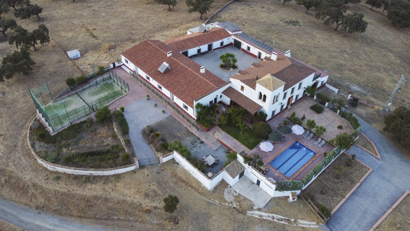 Vista aérea del cortijo: las casas encaladas con tejado de teja alrededor del patio, la piscina, la pista de pádel y la dehesa alrededor